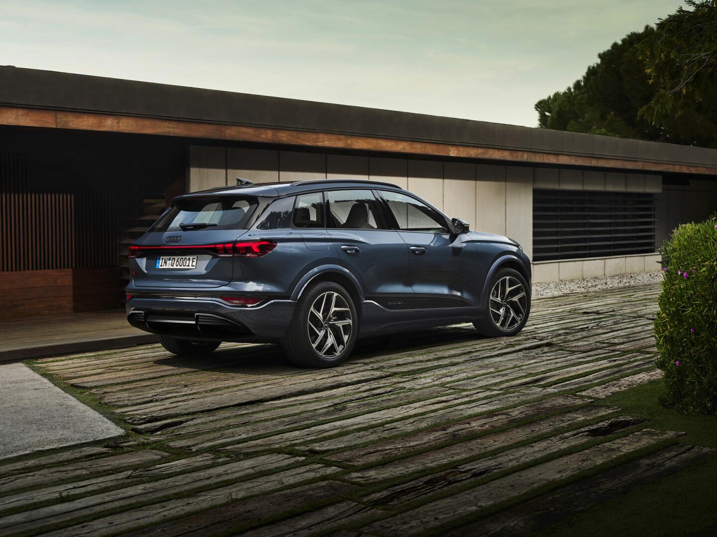 Rear 3 quarter shot a metallic blue Audi Q6 e-tron driving on a wooden desk next to a eco friendly home